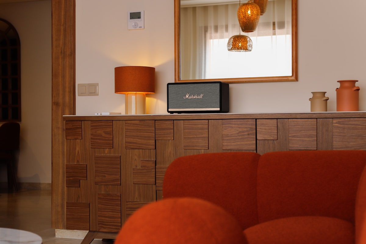A warm living room area features a cozy orange sofa and a stylish wooden sideboard. A Marshall speaker is showcased on the sideboard, accompanied by an amber lamp and decorative vases. Soft lighting creates a relaxed ambiance, complemented by natural light filtering through sheer curtains.