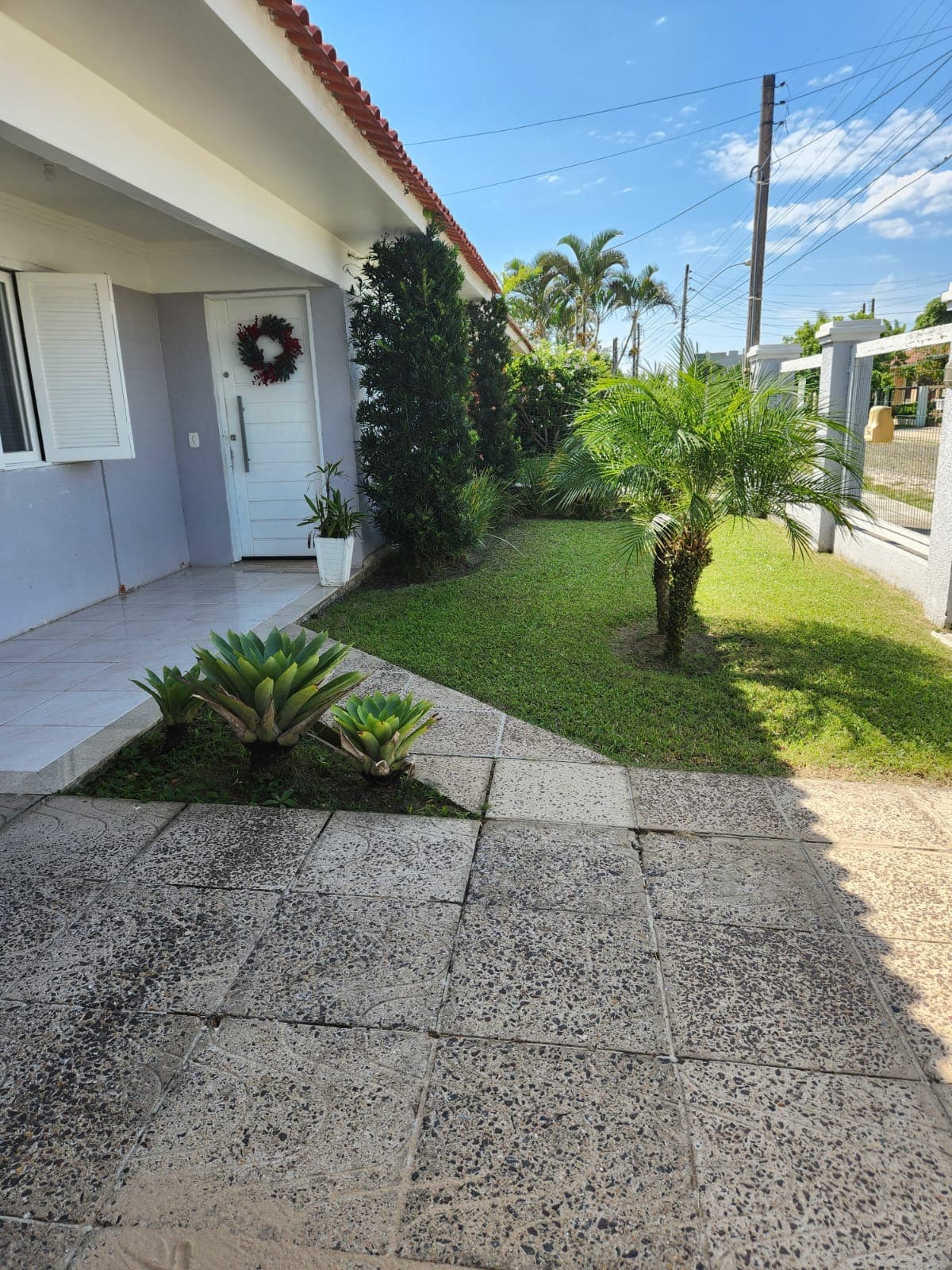 A well-maintained outdoor area features a planted garden with small palms and ornamental plants. A textured stone pathway leads to the entrance of the residence, and a welcoming front door is framed by a festive wreath. Bright sunlight enhances the greenery and neat landscaping.