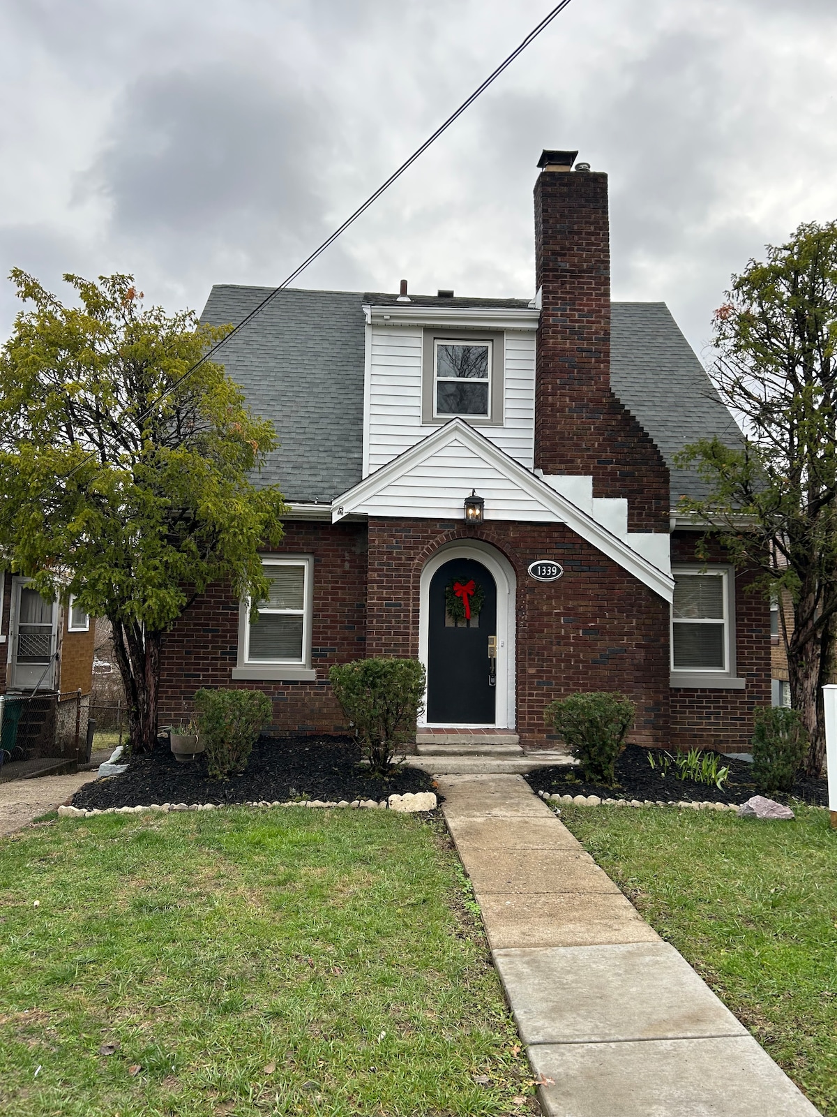 A charming brick house is framed by well-maintained shrubs and small trees. A stone path leads to the front door, which is complemented by a festive wreath. Gray shingles and white accents add to the home's character, set against a cloudy sky.