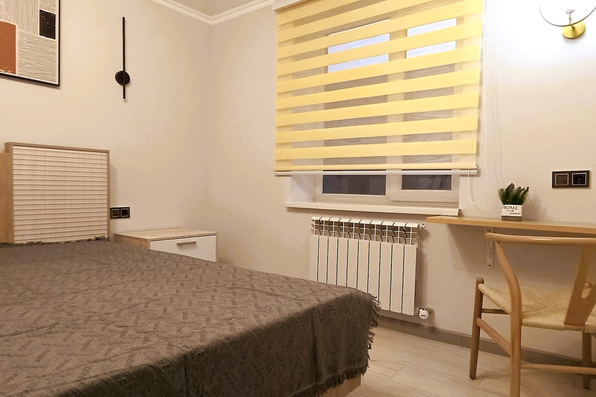 A simple bedroom features a spacious bed with a textured blanket. A yellow striped window covering allows light to filter in. A small desk and chair are positioned near the window, with a potted plant placed on the desk. A radiator is visible on the wall.