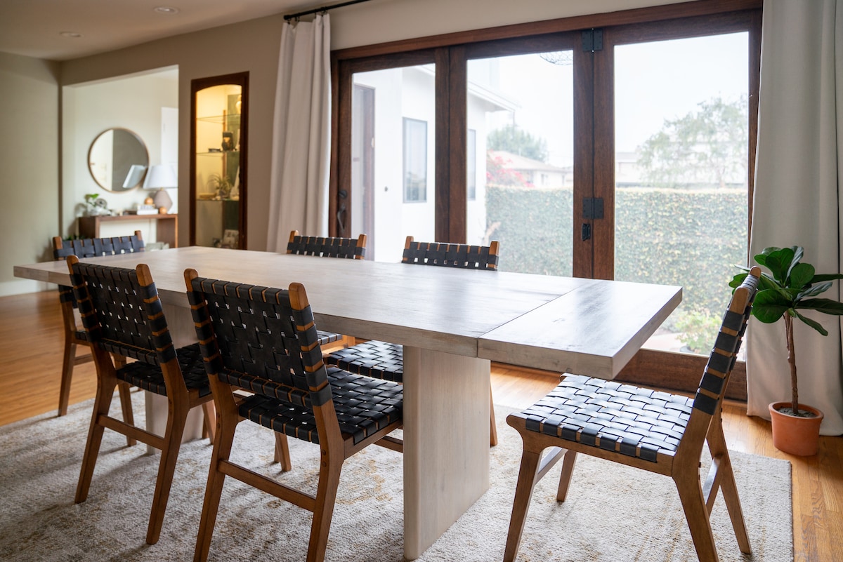 A dining area features a long wooden table surrounded by six woven chairs. Large glass doors provide a view of the outdoor space, while curtains framing the doors offer a soft contrast to the hardwood floors.