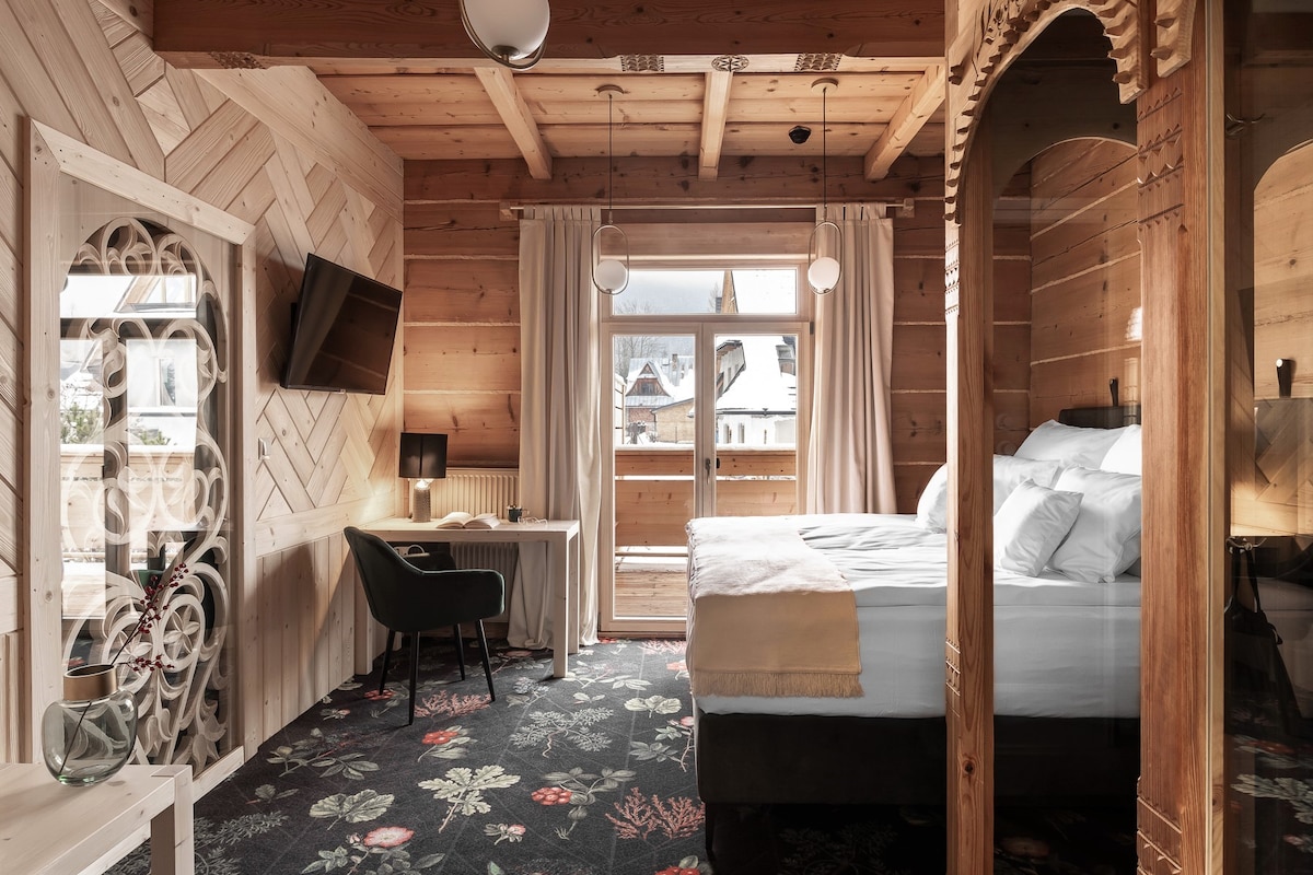 A spacious guestroom features wooden walls and a patterned carpet. A large bed with soft linens is centered in the space. Natural light enters through double doors leading to a balcony, while a TV and a desk with a chair are positioned nearby.