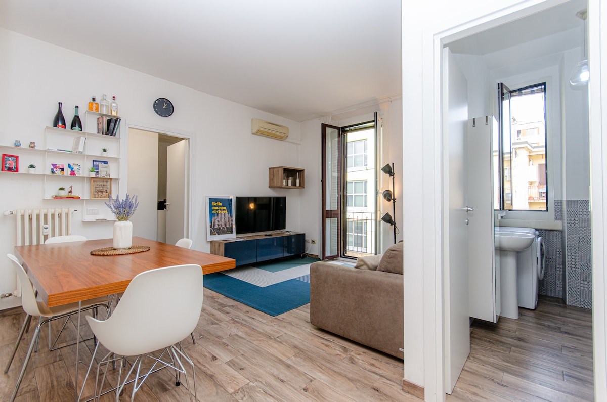 A bright and spacious living area features a wooden dining table surrounded by white chairs, with a flat-screen TV mounted on a modern console. A cozy sofa complements the space. A doorway leads to a well-appointed bathroom, visible in the background.