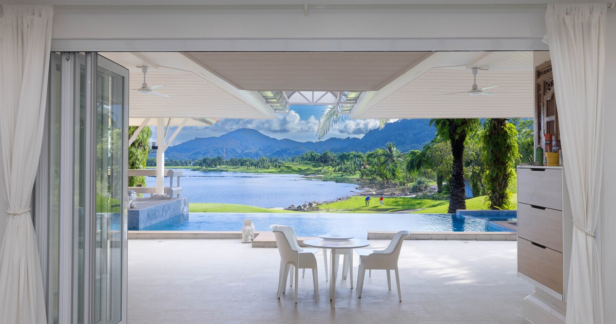 An outdoor dining area features a round table surrounded by four white chairs. The space offers a clear view of a serene lake, palm trees, and rolling hills in the background, creating a calming ambiance. Large open doors invite natural light into the area.