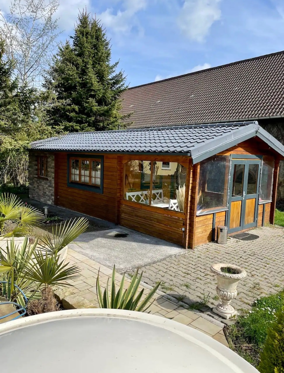 A charming wooden structure is framed by lush greenery and features large windows that invite natural light. The stone pathway leads to a tranquil outdoor area, adorned with potted plants and a small decorative fountain.