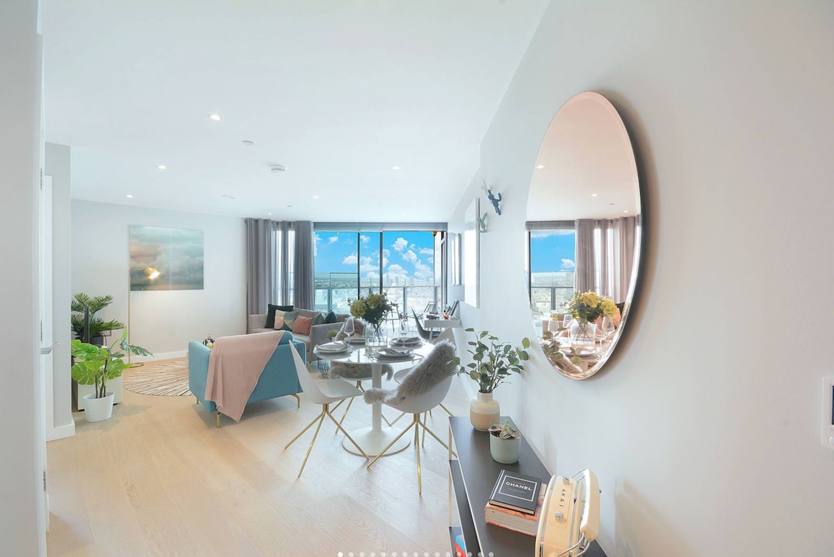 A well-lit living area is shown, featuring a comfortable sofa and a stylish round dining table with chairs. Large windows invite ample natural light and provide a view of the outdoors. A decorative mirror enhances the modern design, complemented by plants and tasteful decor.