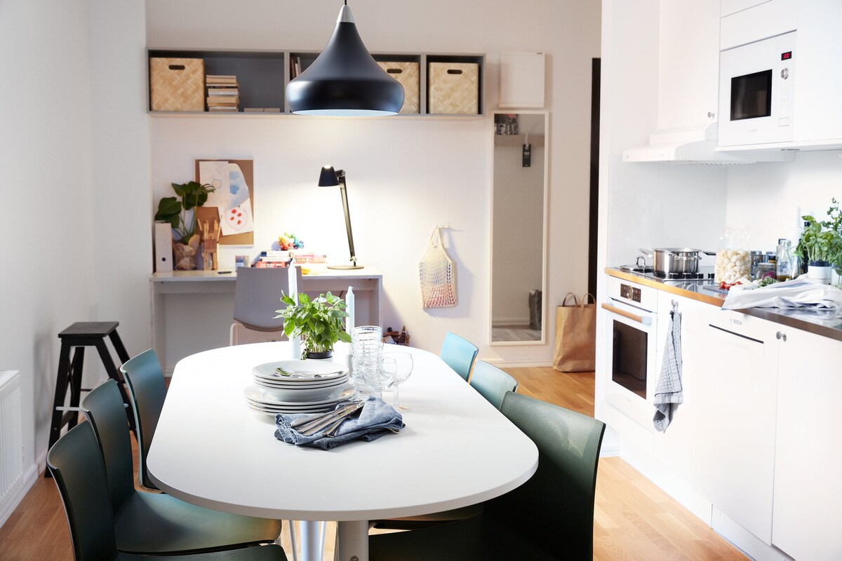 A modern kitchen and dining area are showcased, featuring a round white table with stacked plates and glasses. Behind the table, a stylish pendant light hangs above. A fully equipped kitchen is visible with an oven and cooking elements, while shelves hold neatly arranged baskets and decorative items.