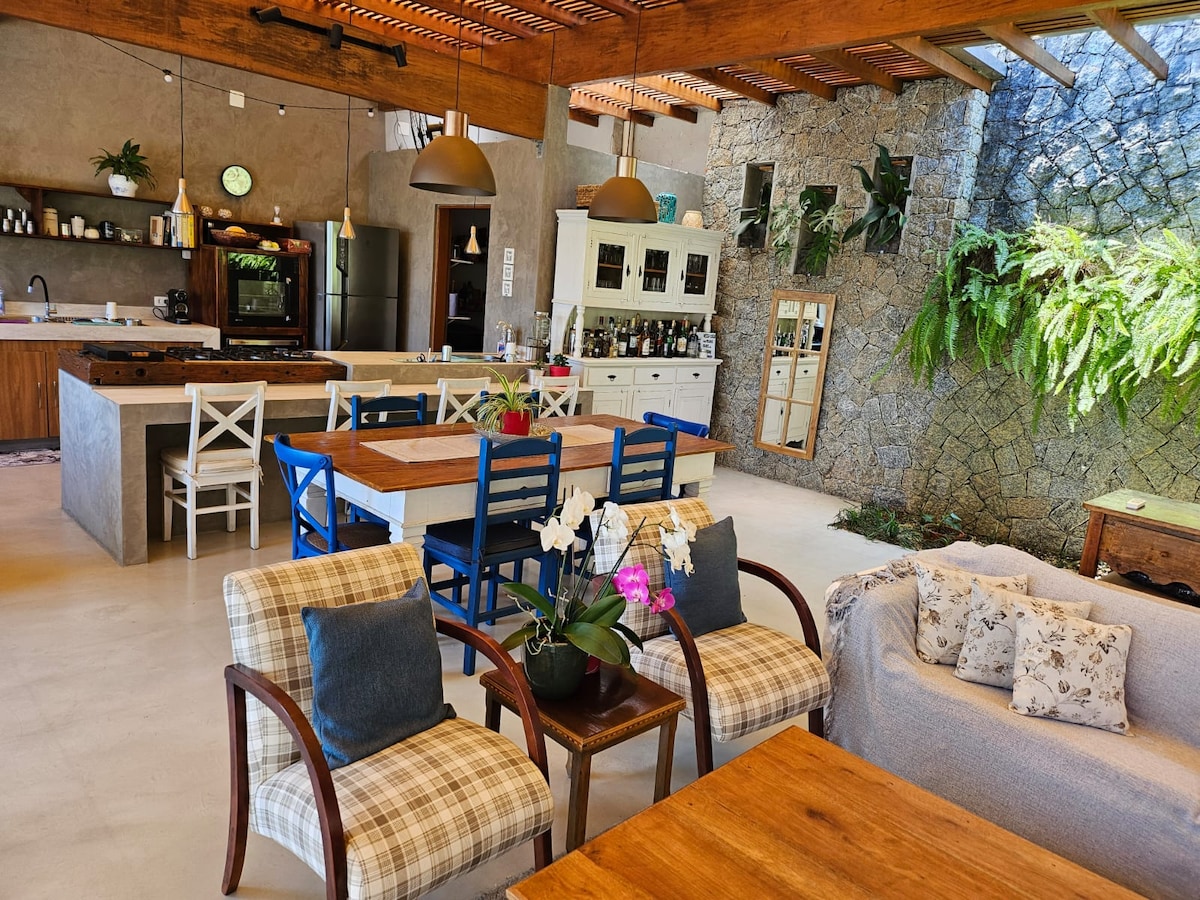 An open-concept kitchen and dining area is highlighted, featuring wooden beams and stone walls. A large dining table accompanied by blue and white chairs is visible, along with cozy seating arrangements. Plants and decorative items enhance the inviting atmosphere of the space.