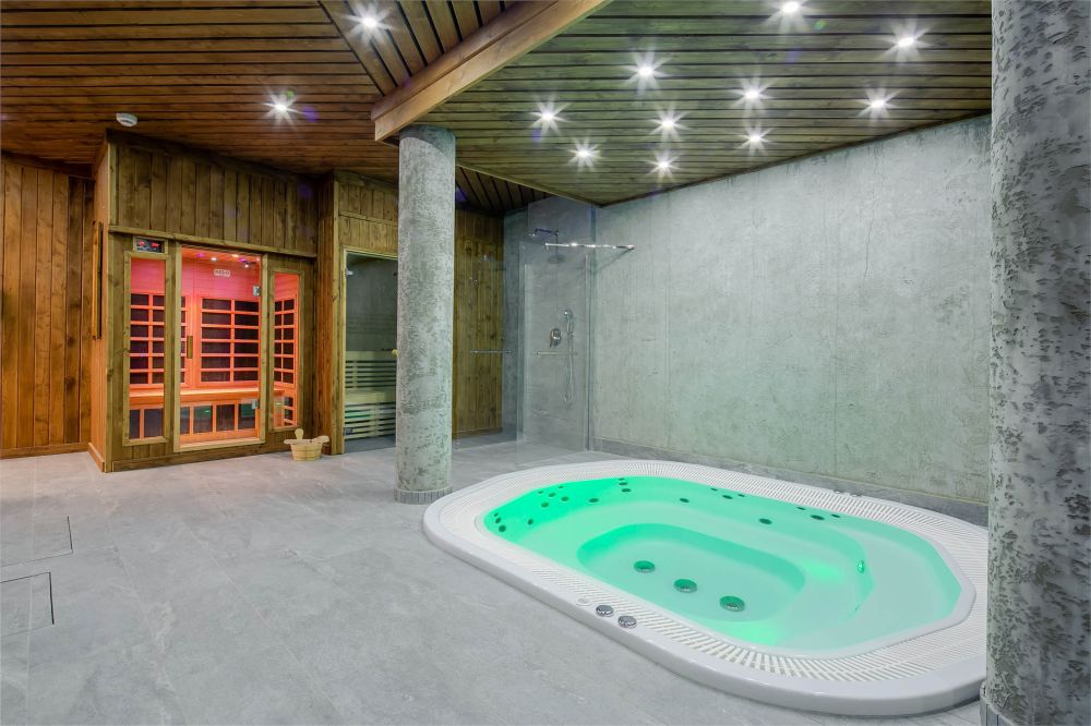A spacious wellness area features a jacuzzi with green lighting, surrounded by textured stone walls. A wooden sauna is visible in the corner, complemented by natural wood beams on the ceiling. Soft, ambient lighting enhances the tranquil setting.