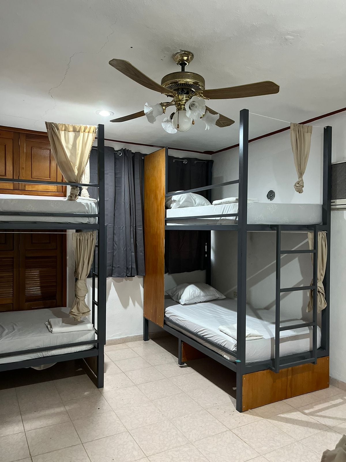 A shared sleeping area is equipped with two sets of bunk beds, featuring comfortable bedding and soft curtains for privacy. The room is illuminated by a ceiling fan and multiple light sources. Neutral tiles cover the floor, while wooden cabinetry adds warmth to the space.