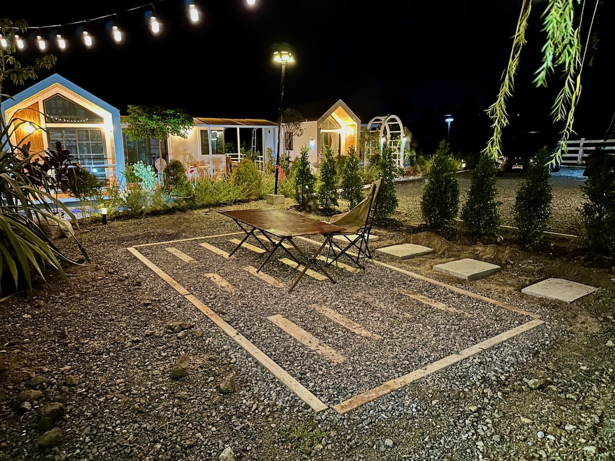 A gravel outdoor area is illuminated by string lights, featuring a cozy seating arrangement with rustic chairs positioned near stepping stones. Small shrubs and carefully arranged plants provide greenery, while charming structures can be seen in the background, enhancing the tranquil night setting.