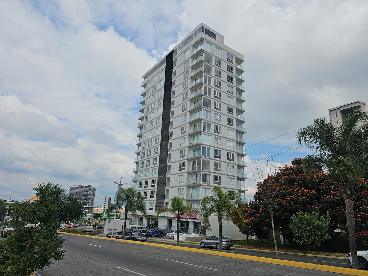 A modern, tall building is pictured along a wide street. Large balconies frame the floors, and greenery is visible in the foreground. Palm trees complement the urban landscape, while the cloudy sky provides a soft backdrop to the structure.