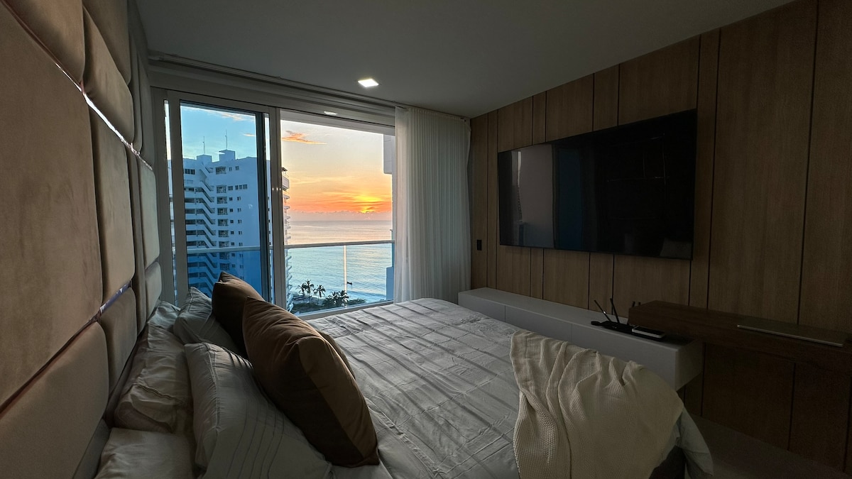 A spacious bedroom is featured with a large bed adorned with neutral-colored linens. A floor-to-ceiling window offers a stunning view of the sunset over the ocean, illuminating the room with warm hues. A flat-screen TV is mounted on the wooden wall opposite the bed.