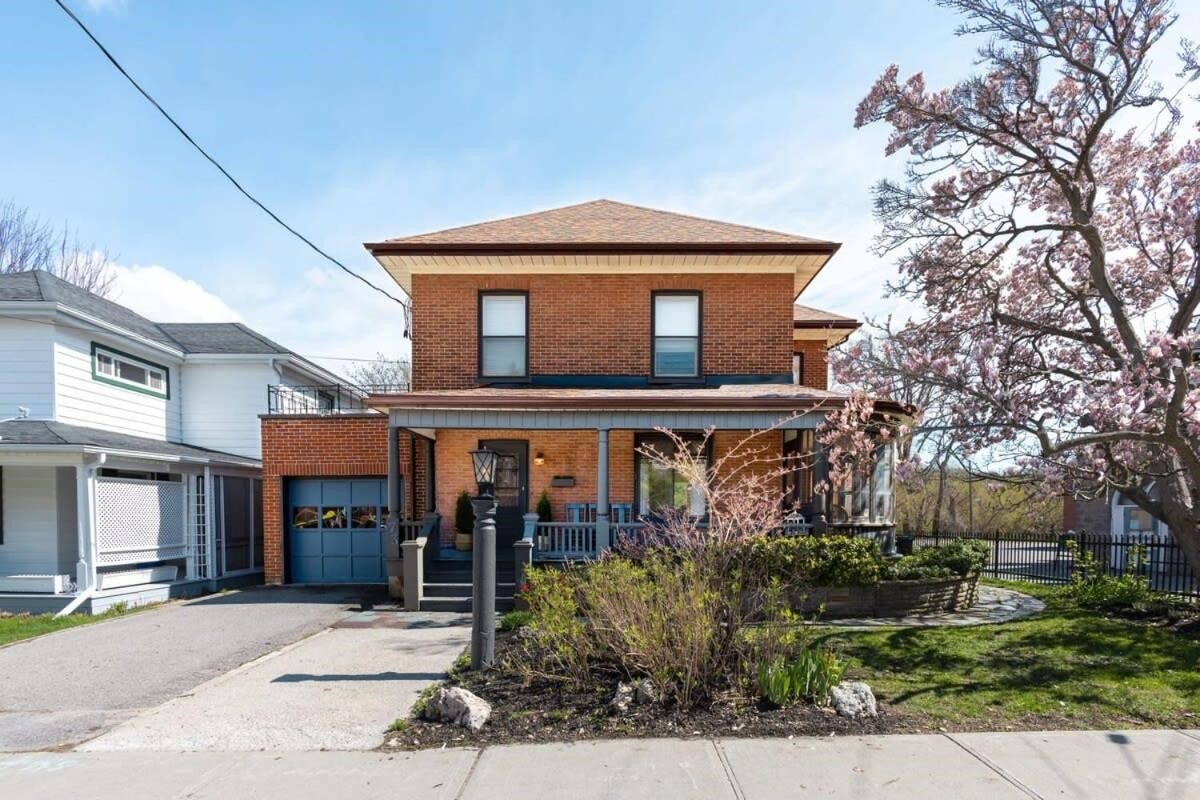 A charming two-story brick house is presented, featuring a warm brown roof and a welcoming front porch. Lush greenery and flowering plants adorn the landscaped yard, and a paved driveway is visible leading to a one-car garage. The clear blue sky provides a bright backdrop.