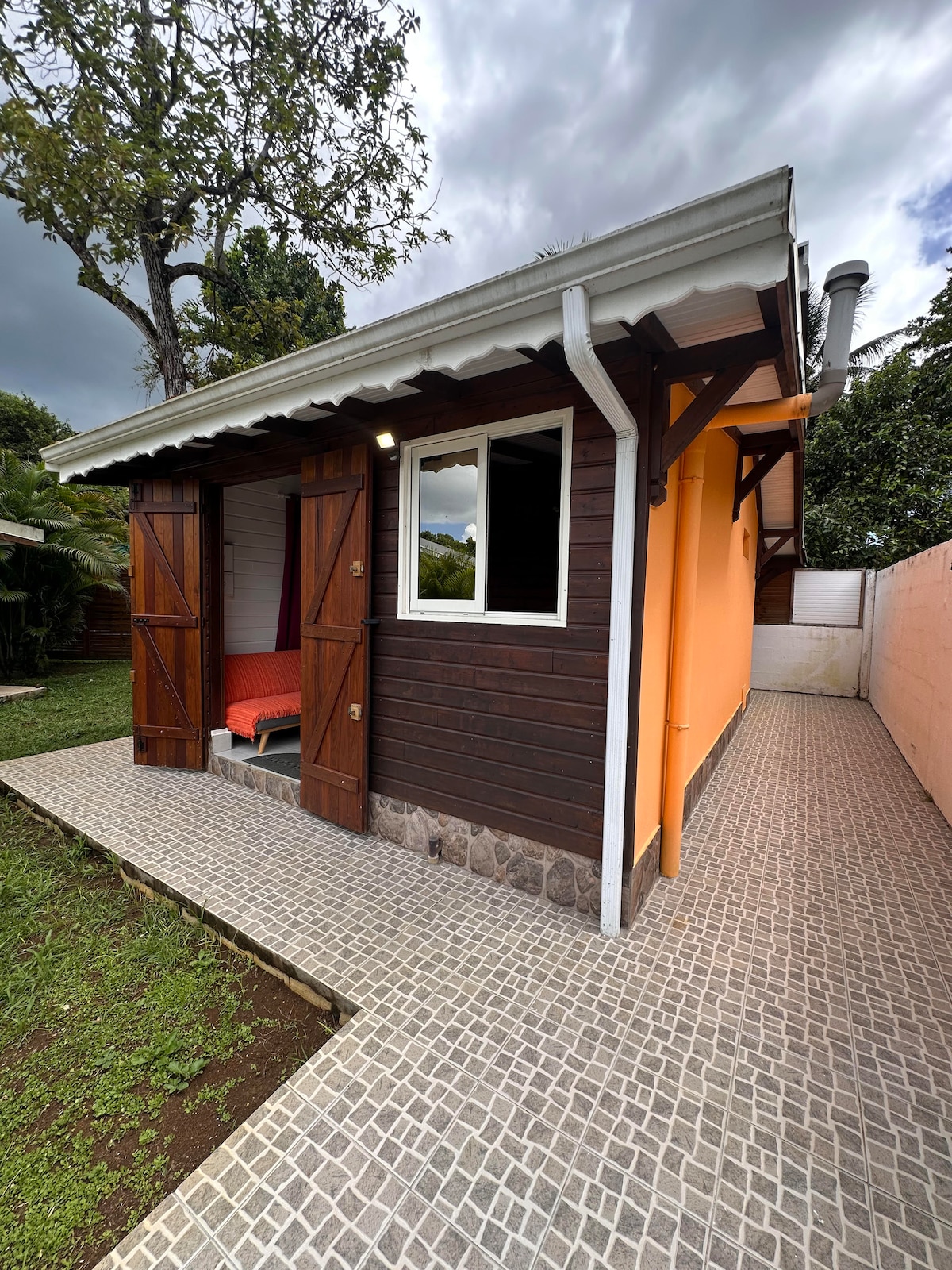 A charming bungalow exterior is presented, featuring wooden double doors that open to a cozy seating area. The building showcases a mix of dark wood and orange siding. A stone pathway leads to a well-maintained lawn, creating a tranquil outdoor space.