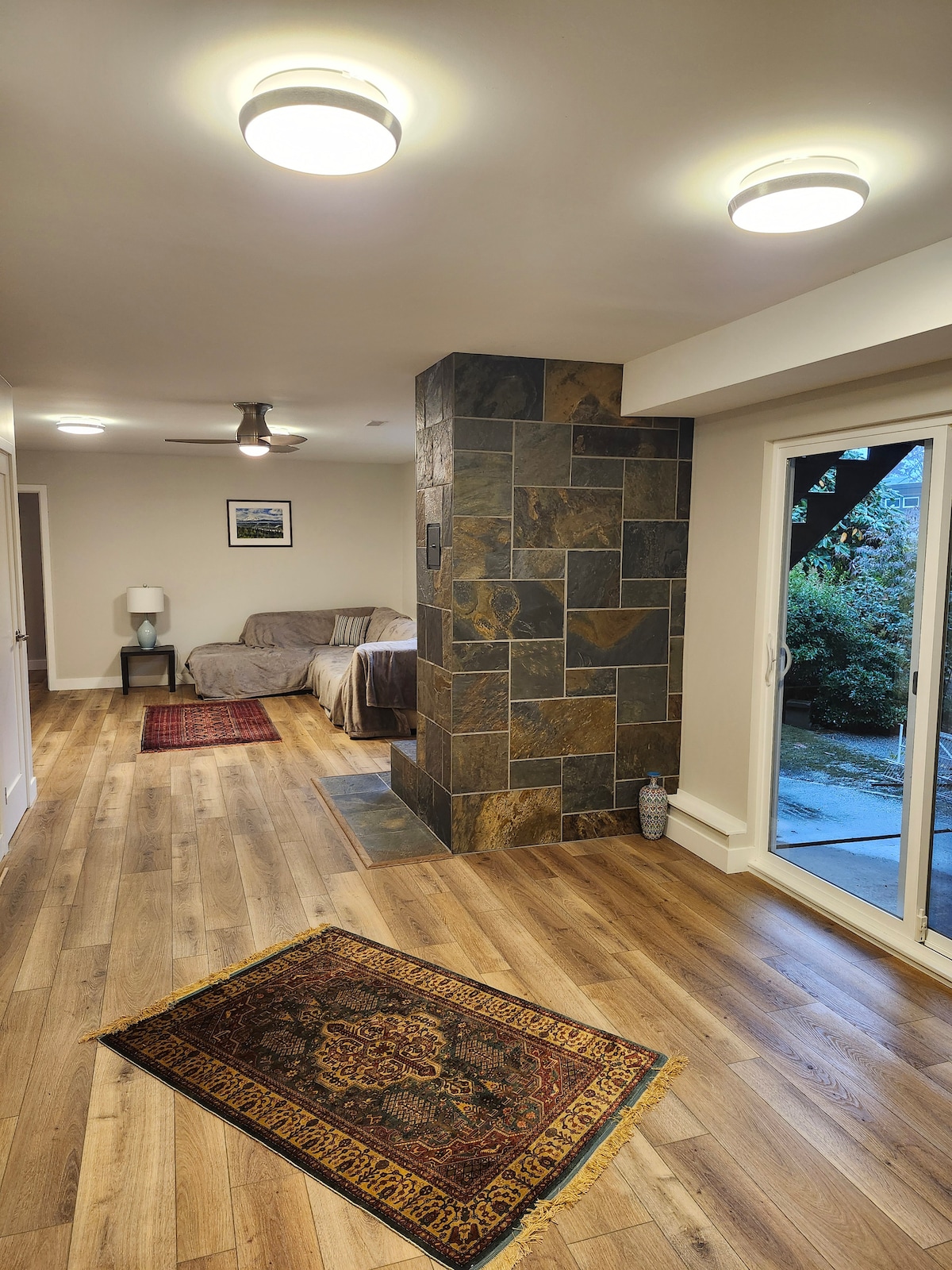 A spacious interior is highlighted by polished wooden floors and a slate stone column. A neutral-toned sofa and a side table are visible in the background, while patterned rugs add warmth. An outdoor patio is accessible through large sliding glass doors.