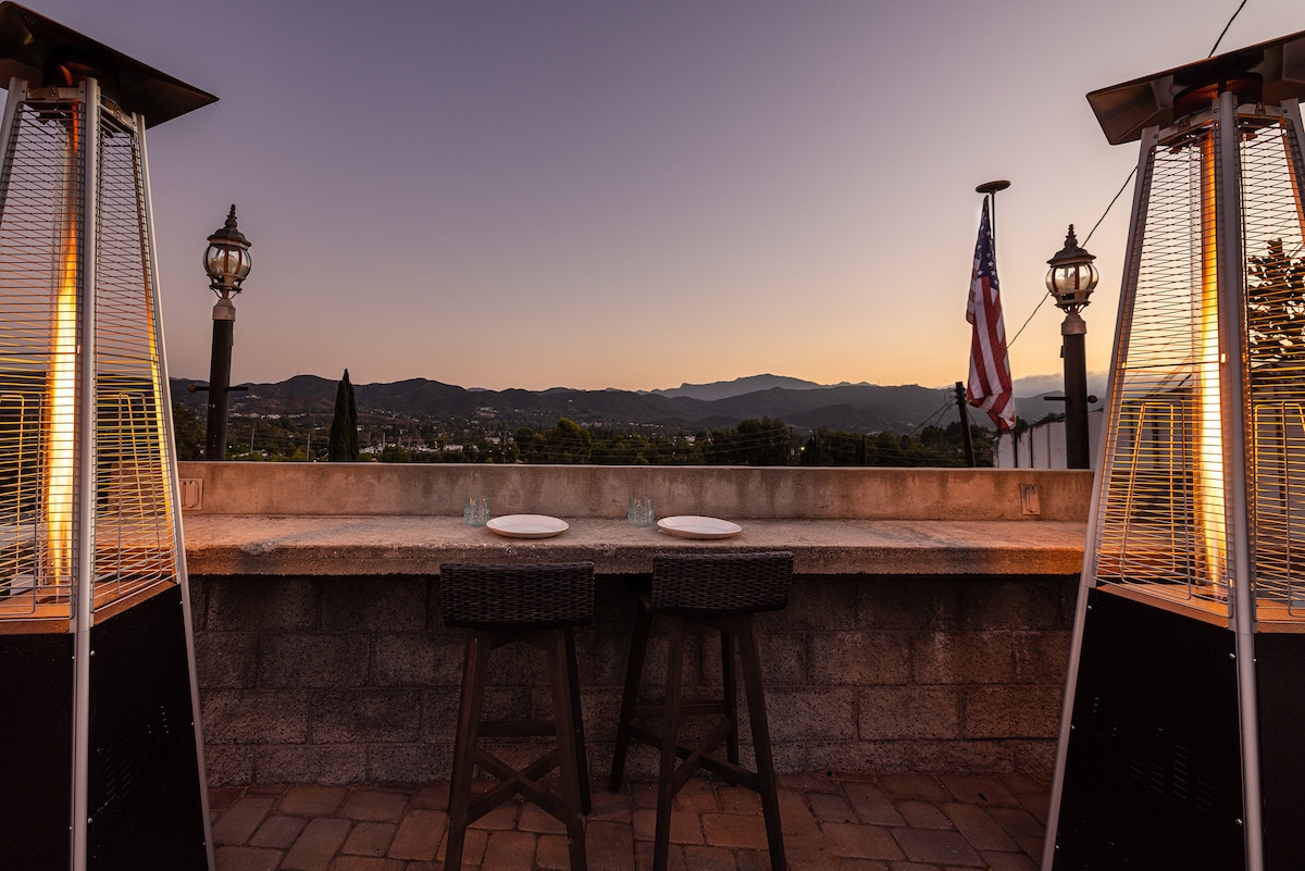 An outdoor dining area is positioned against a backdrop of distant mountains and a clear evening sky. Two tall patio heaters flank a stone table set for two, with white plates and glasses prepared for an al fresco meal. An American flag is visible in the background.
