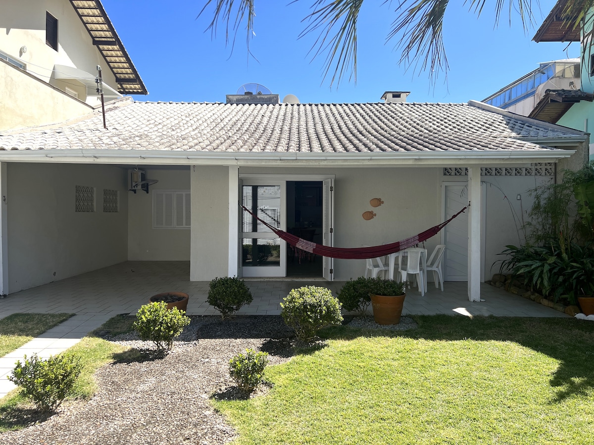 A covered porch with a white exterior features a hammock strung between two posts. A small seating area with white chairs is visible, surrounded by well-maintained shrubs and a patch of grass. Natural light brightens the space, emphasizing the relaxed atmosphere.