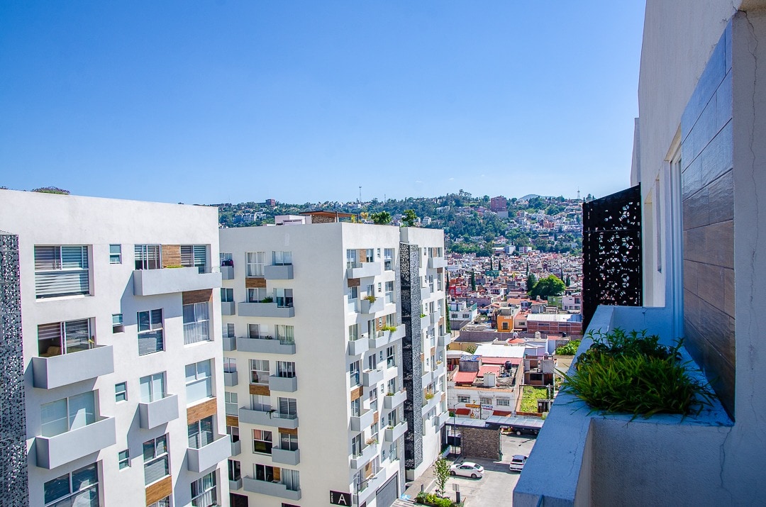 A view from the balcony showcases a modern urban landscape featuring white buildings with varied architectural designs. The backdrop includes a hillside with greenery and gently sloping terrain, hinting at the charm of Morelia's surroundings. Clear blue skies create a bright atmosphere.