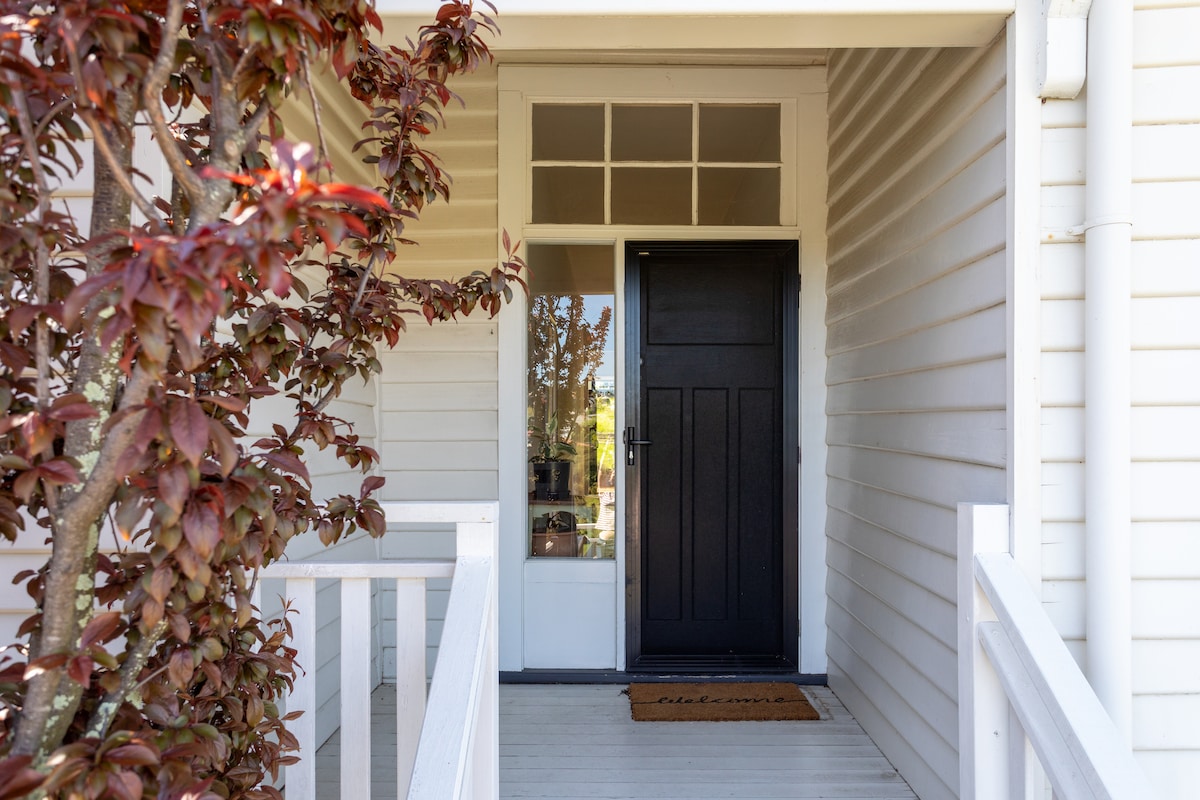 The front entrance features a dark wooden door, framed by large windows that invite natural light. A welcoming doormat lies at the entrance, and colorful foliage from a nearby plant adds a touch of vibrancy to the neutral exterior.