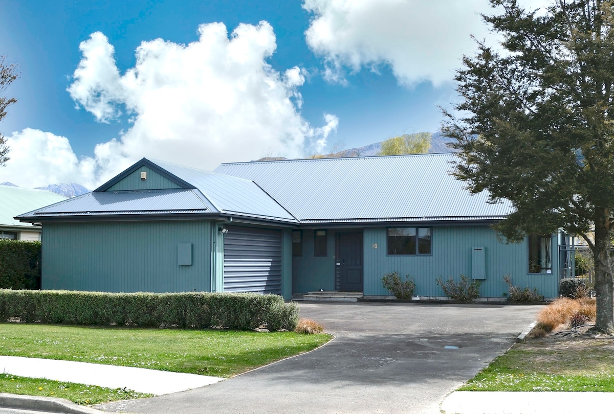 A single-story house is shown with a modern design, featuring a blue exterior and a metal roof. The front yard is landscaped with greenery and hedges. A paved driveway leads to a lockable garage, while the surrounding area includes a clear sky with scattered clouds.