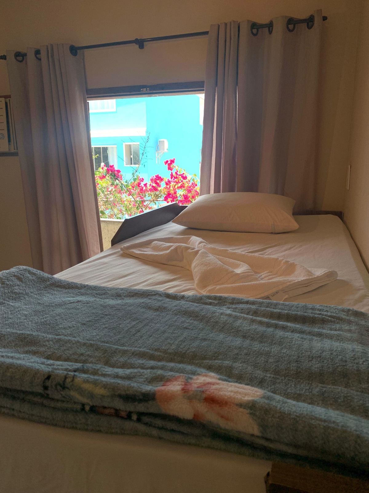 A simple sleeping area features a single bed dressed with a white sheet and light-colored pillow. A blue blanket is casually placed at the foot of the bed. Sunlight enters through a window adorned with curtains, revealing vibrant flowers and a colorful building outside.
