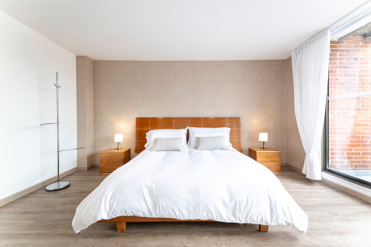 A spacious bedroom features a comfortable queen-sized bed dressed in crisp white linens. Two bedside lamps provide soft lighting, and large windows allow natural light to illuminate the room, showcasing the warm wood accents on the headboard and side tables.