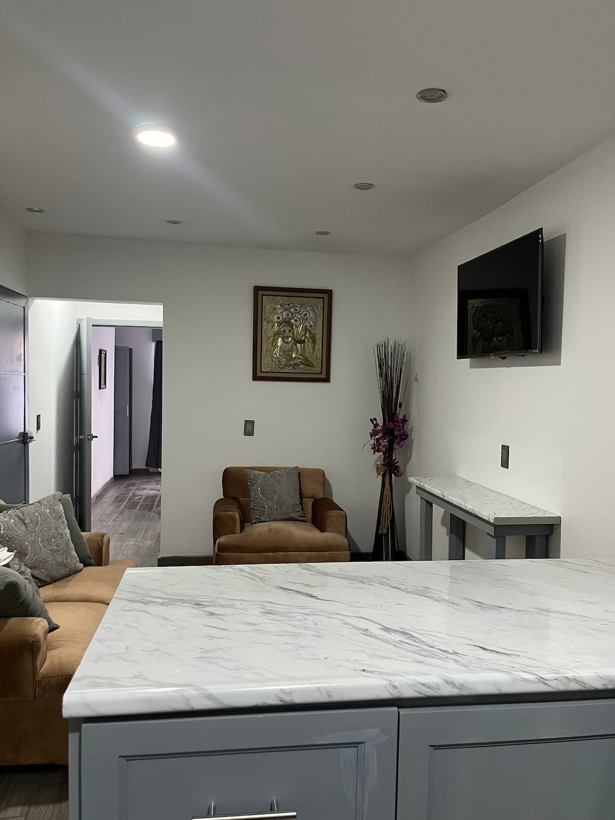 A modern living area features two comfortable brown armchairs and a marble-topped table. A framed piece of wall art is displayed, alongside a flat-screen TV mounted on the wall. The open doorway leads to additional living space, enhancing the room's connectivity.