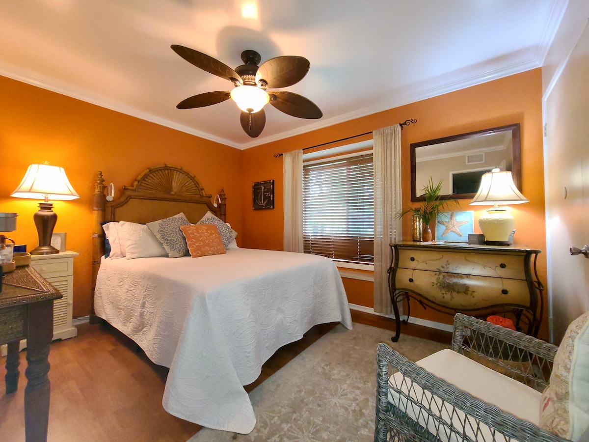 A warm orange room features a spacious bed with a light quilt and decorative pillows. Two lamps illuminate a wooden nightstand on one side, while a vintage dresser is positioned across from a cozy woven chair. Natural light filters through the window, enhancing the inviting ambiance.