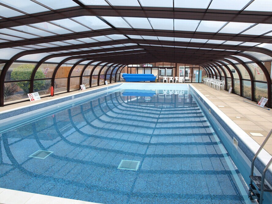 An indoor pool is enclosed by a transparent roof, allowing natural light to illuminate the water's surface. Surrounding the pool are tiled walkways and safety signage. A blue inflatable pool toy floats on one side, with several white chairs positioned nearby.