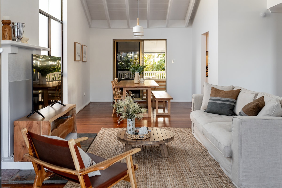 The open-plan living area features high ceilings and large windows allowing natural light to enter. A comfortable sofa is arranged around a circular coffee table, with an armchair nearby. A dining table is visible in the background, promoting a welcoming atmosphere for gatherings.