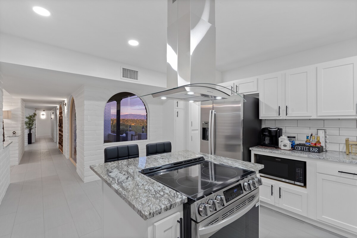 A modern kitchen features a central island with a granite countertop, equipped with seating for two. Stainless steel appliances and white cabinetry provide a clean and bright appearance. Natural light enters through an adjoining window, highlighting the sleek design and spacious layout.