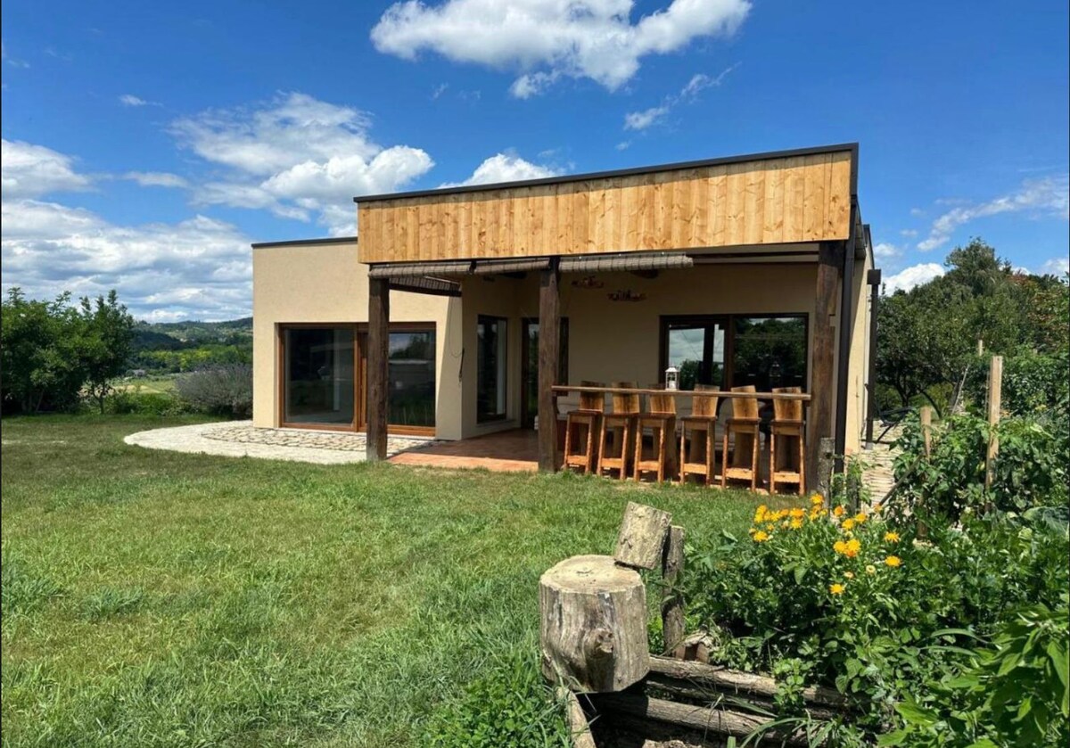 The exterior of the apartment features a modern design with a wooden overhang and large windows, allowing plenty of natural light. A spacious patio area includes a row of wooden bar stools, overlooking a grassy landscape adorned with flowers and trees.