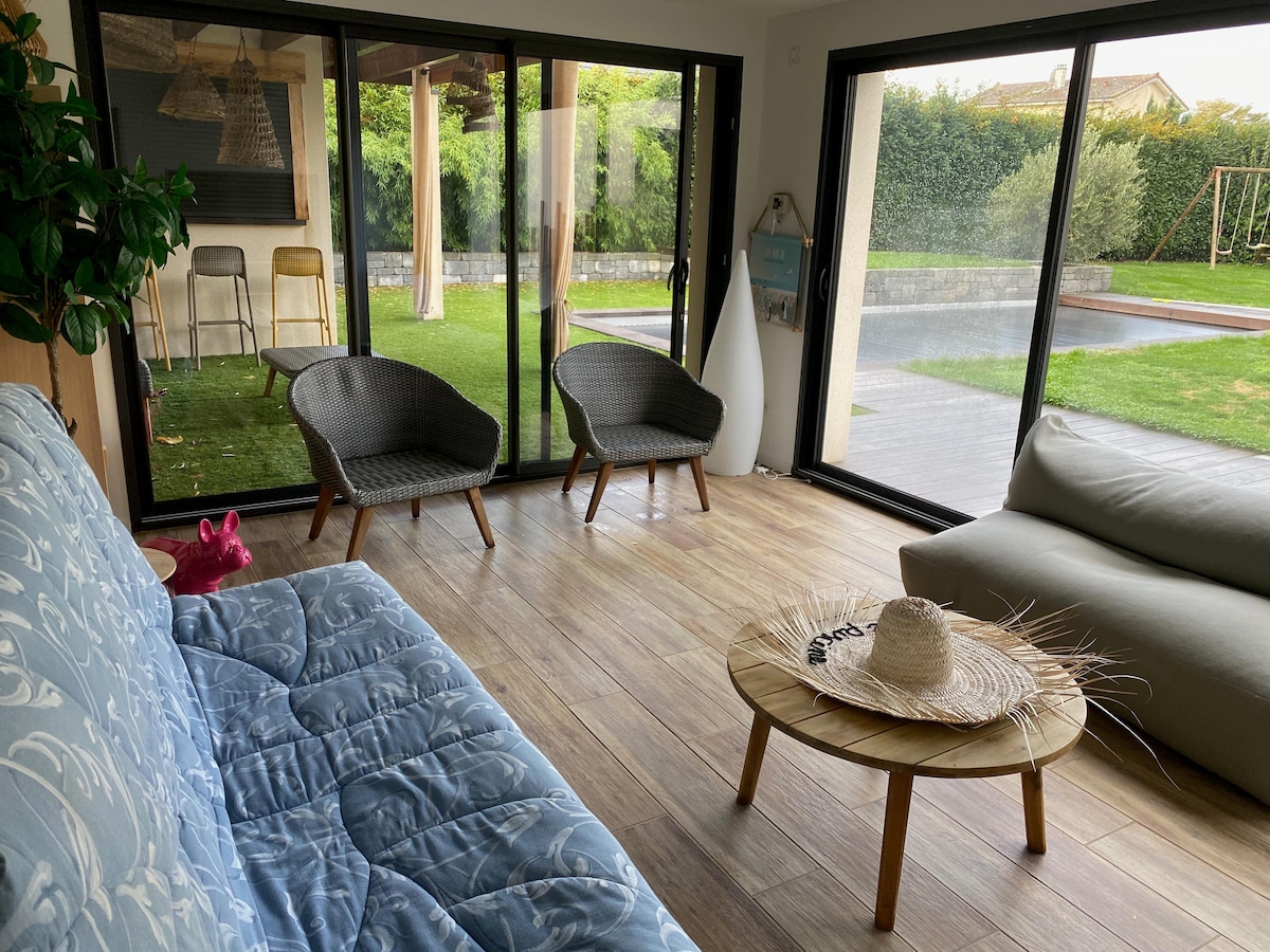 A relaxed seating area features two gray armchairs and a soft light-colored sofa. A round wooden coffee table sits in front, adorned with a straw hat. Large glass doors open to a green outdoor space, showcasing a swimming pool and a swing set.