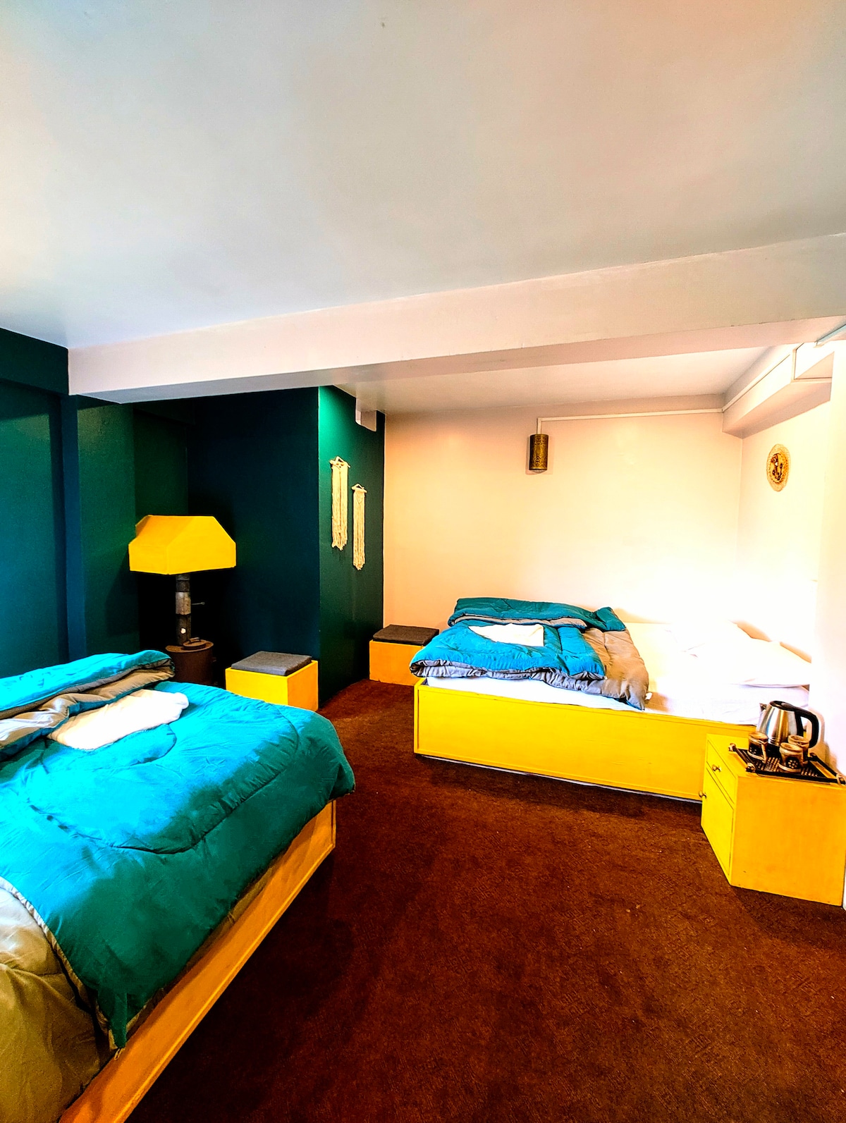 A brightly colored room features two beds with plush bedding, one in vivid teal and another in neutral tones. A lamp stands next to the teal bed, complementing wooden nightstands. The walls are painted in a soft teal and white, creating a cozy and inviting ambiance.