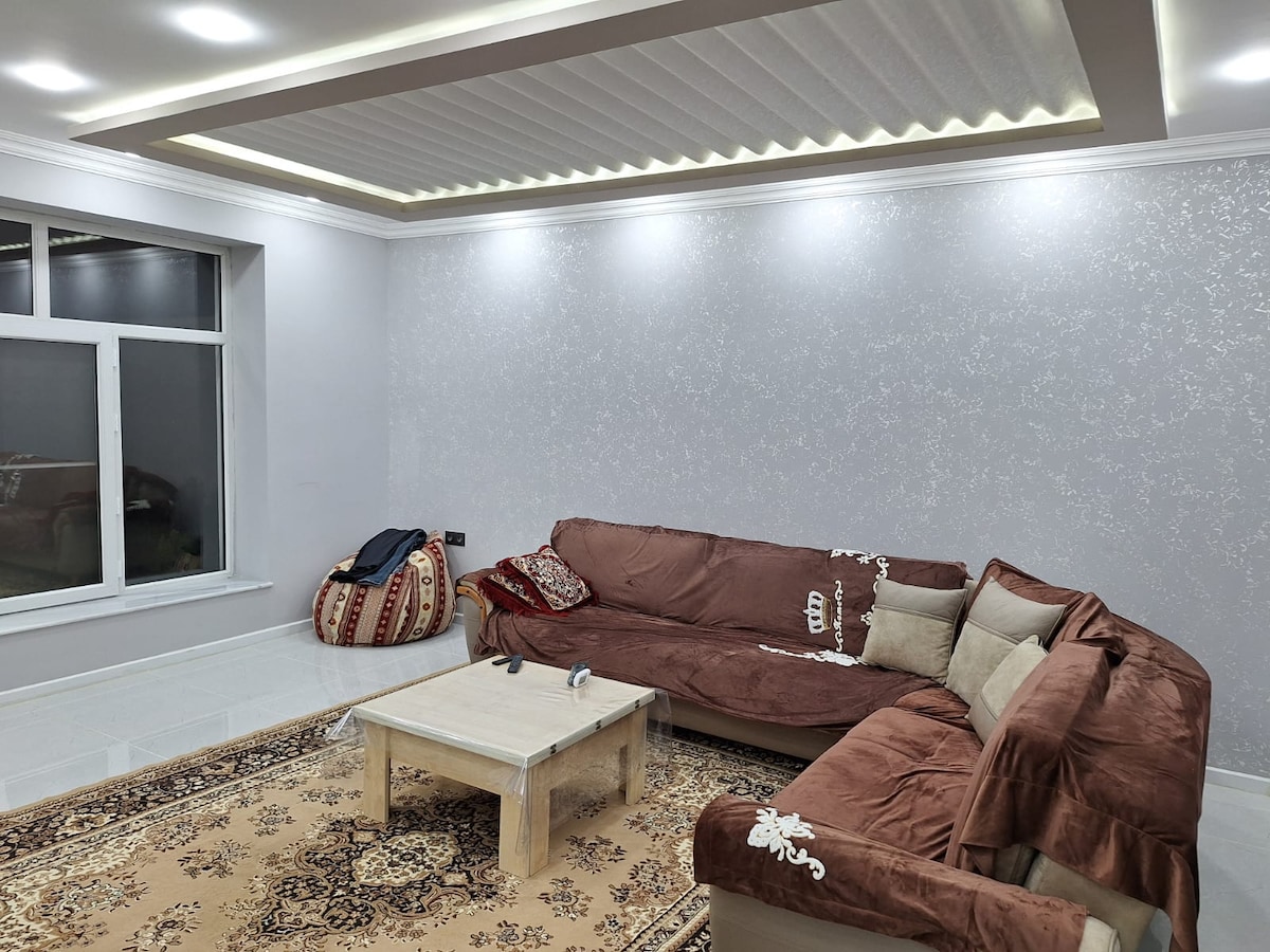 A spacious living area features a light-colored sectional sofa adorned with decorative pillows, positioned around a low coffee table. A large window allows natural light to illuminate the room, complemented by modern ceiling lighting. A patterned area rug adds warmth to the sleek, tiled floor.