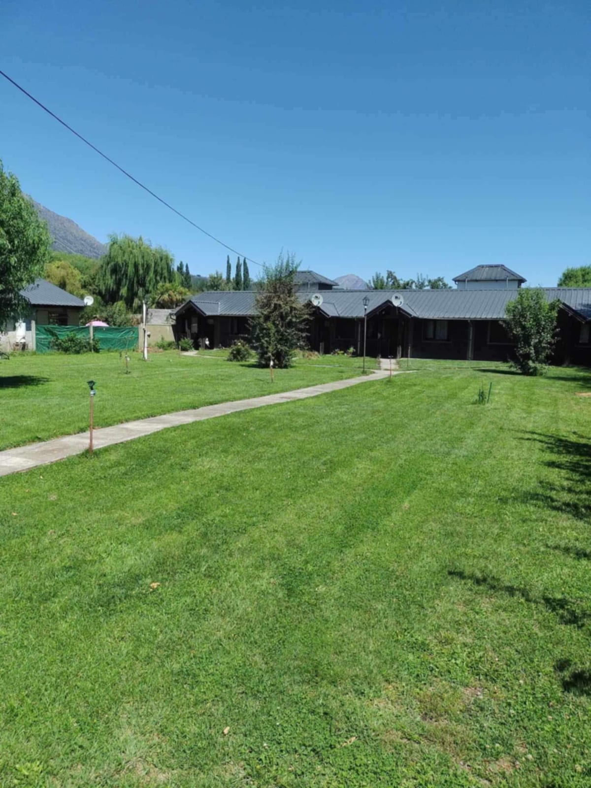 A well-maintained grassy area is presented, with a pathway leading to a series of charming structures nestled among trees and shrubs. A clear blue sky fills the background, enhancing the serene outdoor setting.