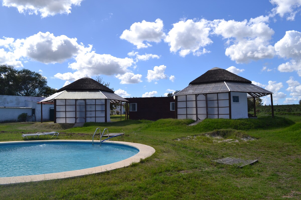 Two bioconstruction cabins with thatched roofs are pictured beside a circular swimming pool, surrounded by green grass. A clear blue sky with fluffy clouds adds to the serene environment, while the pool features a simple ladder for access.