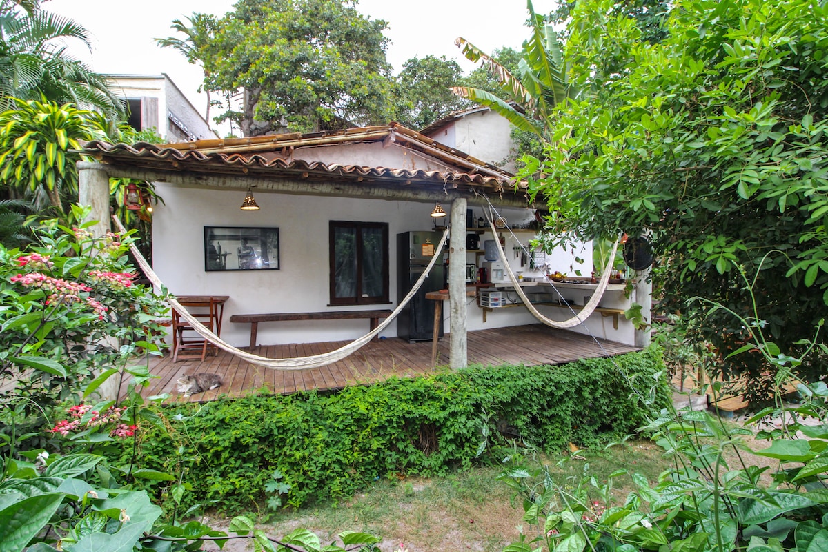 A charming exterior of the house is framed by lush greenery and tropical plants. A wooden deck features a hammock, inviting relaxation. The rustic roof and simple architecture blend with the surrounding nature, showcasing a welcoming space for leisurely moments.