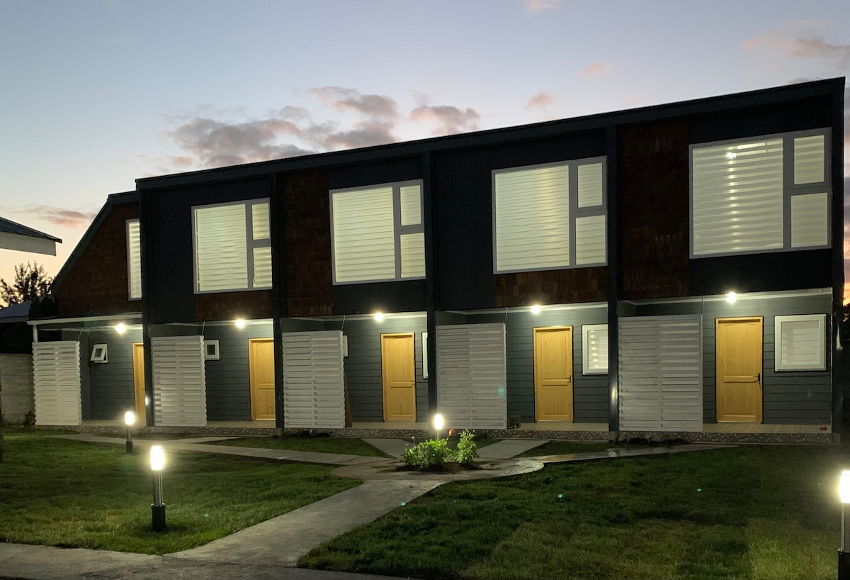 A row of modern accommodations is seen, featuring multiple entrances with illuminated doors. Large windows allow natural light to enter, and well-maintained grass surrounds the entrance pathways, completing the inviting exterior of the building.
