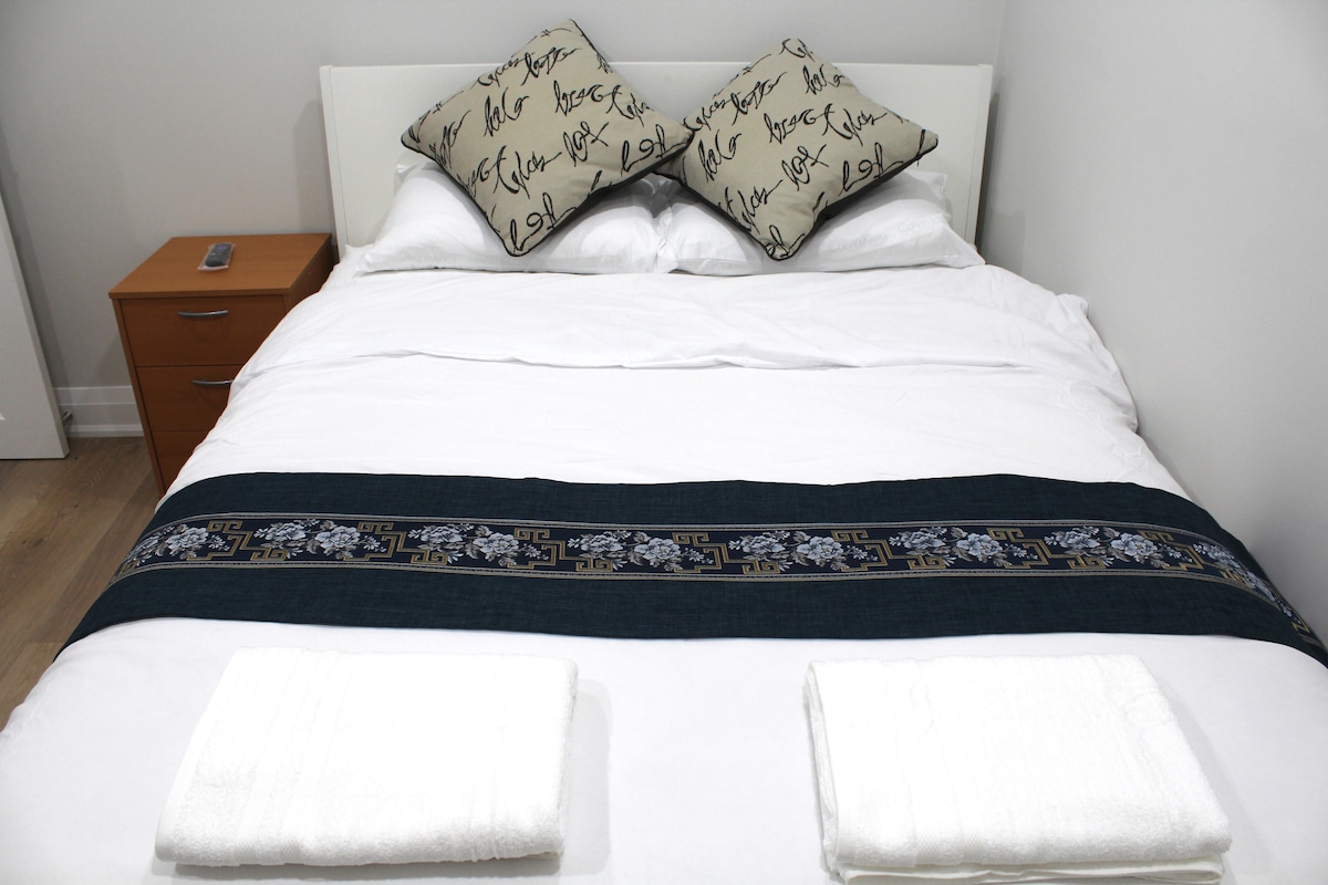 A spacious bed is featured with a dark decorative runner, complemented by two patterned pillows. Fresh white towels are neatly placed at the foot of the bed. A bedside table is visible, equipped with a remote control.