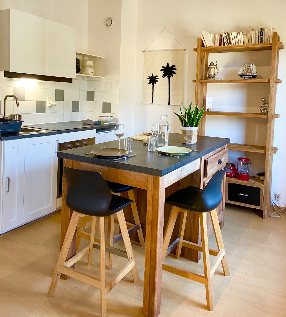 A functional kitchen area is presented, featuring a small wooden dining table with four chairs. A dark countertop complements white cabinets, while a shelf displays decorative items. A plant adds a touch of greenery beside a window with light entering the space.