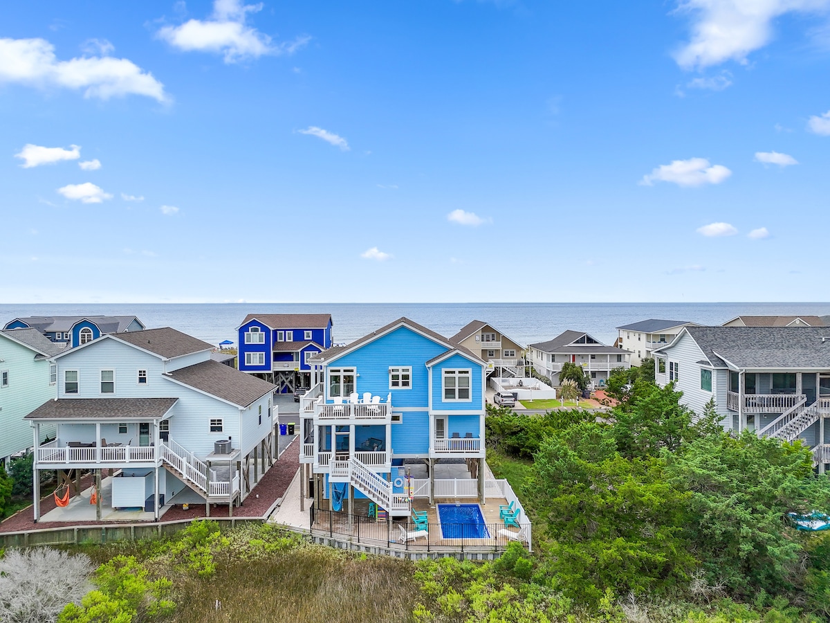 A spacious two-story home is showcased against a backdrop of the ocean. The exterior features a vibrant blue color, with decks overlooking the water. Surrounding homes are visible, set amidst greenery and sandy paths leading to the beach.