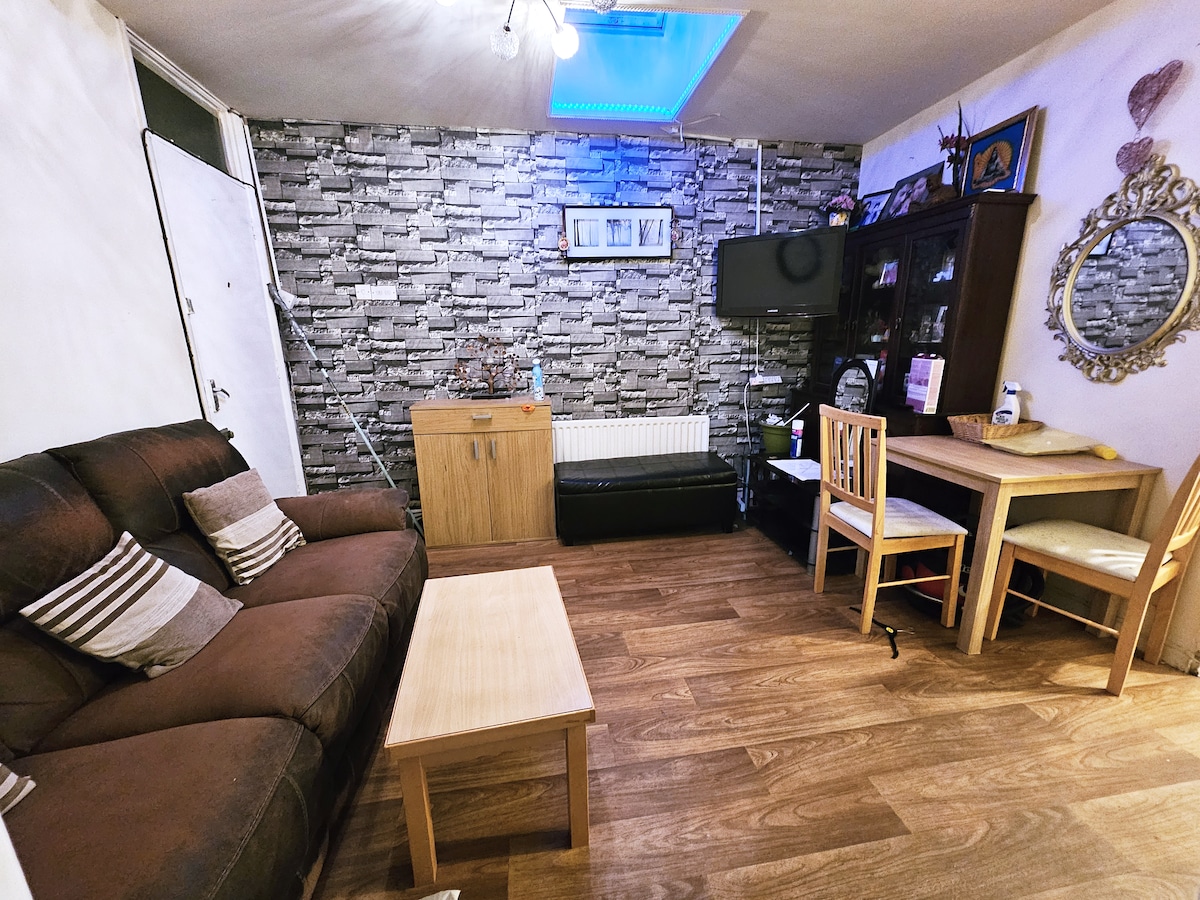 The living room features a comfortable brown sofa with decorative cushions, a wooden coffee table, and a small dining area with two chairs. A stylish cabinet holds various items, and a television is mounted on the wall. Natural light is provided by a skylight.