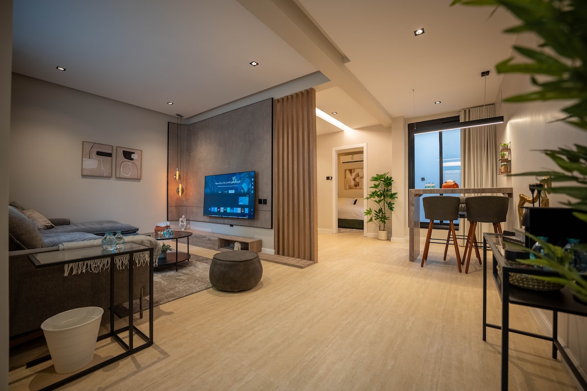 A spacious living area is equipped with a modern television mounted on a neutral-colored wall. A comfortable seating arrangement features a dark sofa, a coffee table, and natural light streaming through large windows. The dining space is visible, showcasing stylish chairs and greenery.