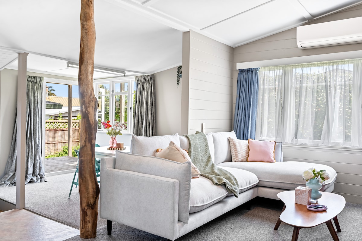 A bright lounge area features a spacious, light-colored sofa accompanied by soft pillows and a textured throw. Large windows invite natural light and provide views of the outdoor deck. A small side table holds a vase of flowers, enhancing the cozy setting.