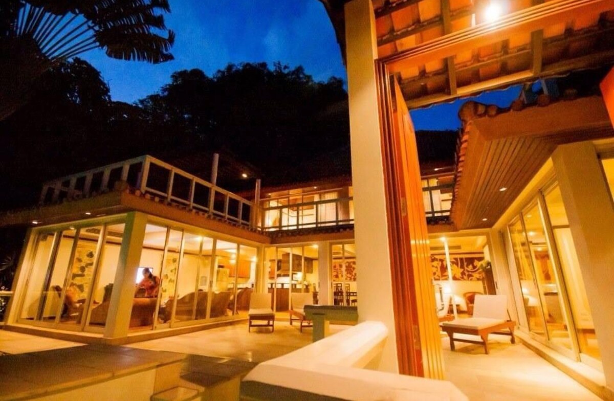 The villa's exterior is illuminated at dusk, showcasing modern architecture with expansive glass walls. The inviting outdoor area features comfortable seating and a view of lush greenery, creating a seamless connection between indoor and outdoor living spaces.