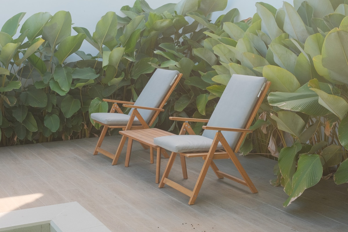 Two comfortably cushioned lounge chairs are positioned on a wooden deck, surrounded by lush green foliage. The natural setting provides a peaceful spot for relaxation, ideal for enjoying the outdoors.