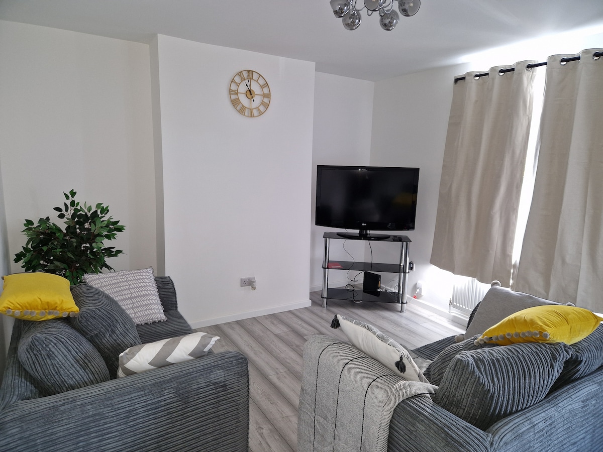 A cozy sitting area features two gray sofas adorned with yellow and striped cushions. A sleek TV sits on a stand, complemented by light curtains that frame the window. A small decorative plant adds a touch of greenery, enhancing the inviting space.