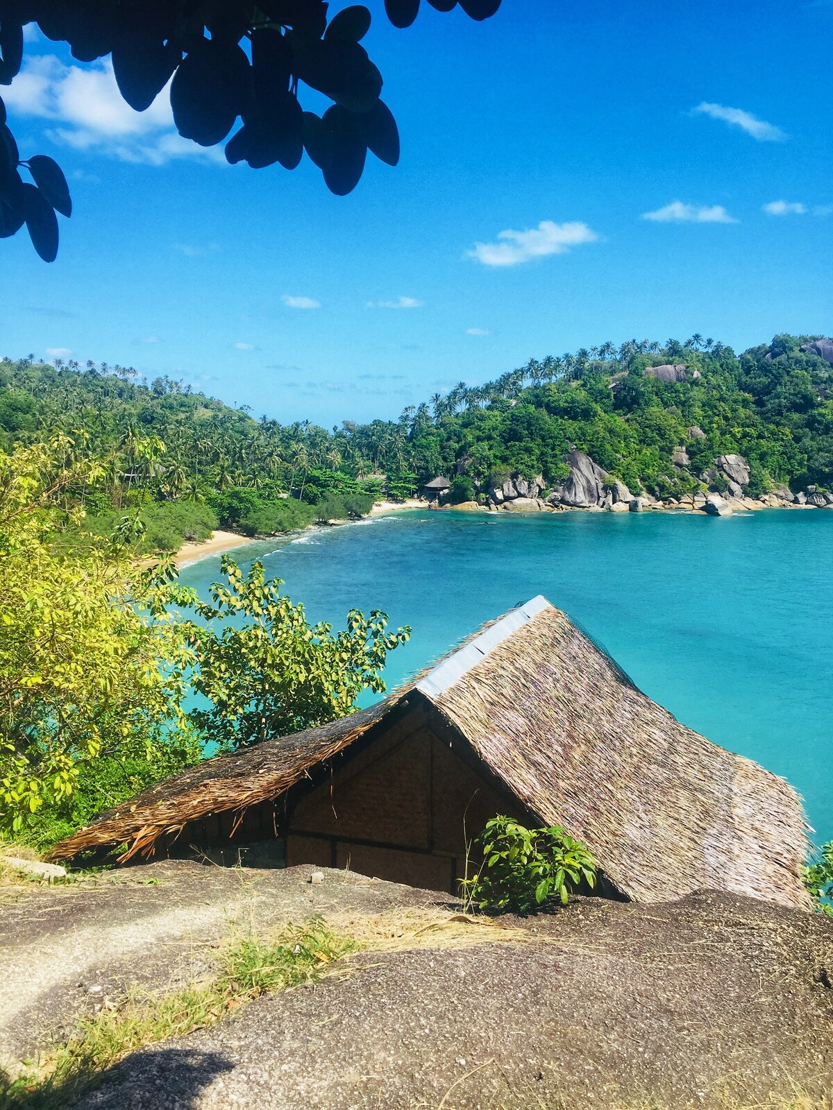 A thatched-roof bungalow is nestled on a rocky outcrop, surrounded by lush greenery. The vibrant turquoise water of the bay glimmers in the sunlight, while hills and rock formations frame the serene seascape in the background.